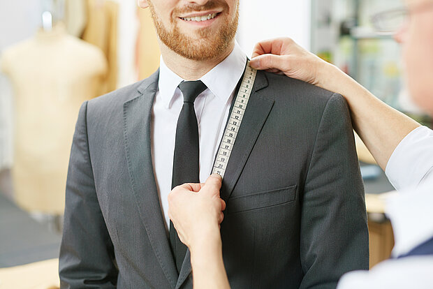 Ein Mann in einem Anzug schaut zum Schneider und lacht. Der Schneider hält ein Maßband in der Hand und misst die Größe seiner Jacke von Schulter bis Brust.