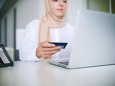 Eine Frau mit Kopftuch sitzt vor ihrem Laptop und hält eine Karte in der Hand.