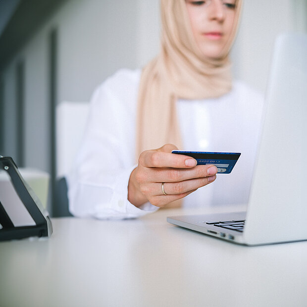 Eine Frau mit Kopftuch sitzt vor ihrem Laptop und hält eine Karte in der Hand.