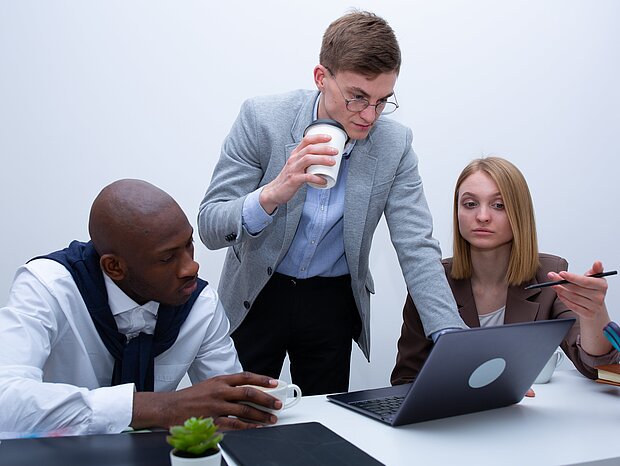 Eine Frau und zwei Männer sitzen am Schreibtisch, auf dem ein Laptop, ein iPad und Ordner liegen, und besprechen ein Thema