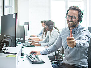Im Büro sind mehrere Mitarbeiter zu sehen. Ein fröhlicher Mitarbeiter im Vordergrund trägt ein Headset, lacht, zeigt den Daumen nach oben und hat eine Brille auf.