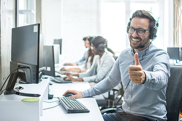 Im Büro sind mehrere Mitarbeiter zu sehen. Ein fröhlicher Mitarbeiter im Vordergrund trägt ein Headset, lacht, zeigt den Daumen nach oben und hat eine Brille auf.