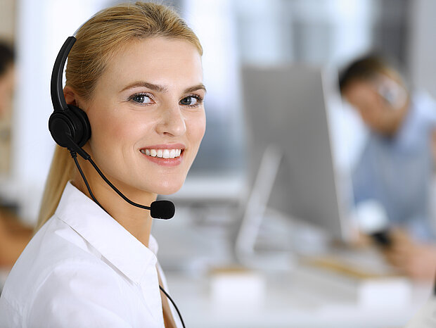 Eine blonde Frau in einem weißen Hemd und mit einem Headset sitzt im Büro