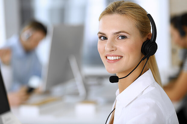 Eine blonde Frau in einem weißen Hemd und mit einem Headset sitzt im Büro