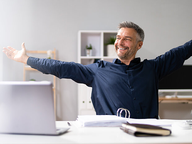 Ein Mann sitzt im Büro mit erhobenen Händen, lacht und freut sich. Vor ihm auf dem Tisch befinden sich ein PC, Laptop, Ordner, ein Heft und eine Brille.