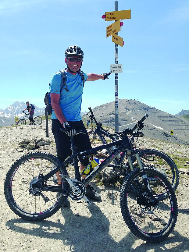 Ein Mann mit Helm und Fahrrad steht neben einem Schild. Das Schild enthält verschiedene Ortsnamen und Wegweiser.