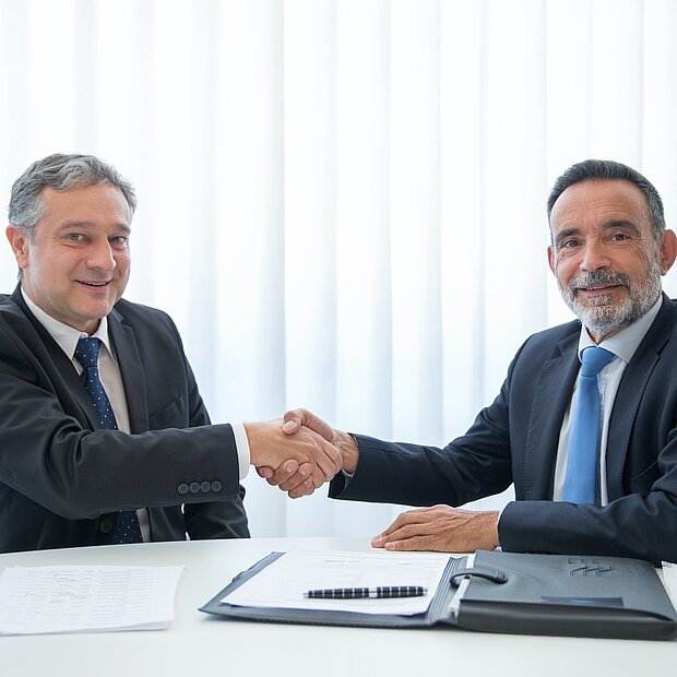 Zwei Männer in Anzügen sitzen an einem Tisch, auf dem ein Vertrag liegt, und geben sich die Hand