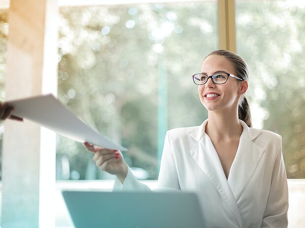 Eine Frau mit weißem Anzug und Brille sitzt im Büro und gibt Unterlagen an eine andere Person weiter. Vor ihr liegt ihr Laptop.