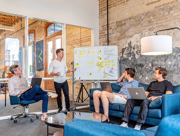 Vier Männer sind im Teammeeting, während einer am Whiteboard präsentiert. Auf der Tafel sind mehrere Notizzettel.