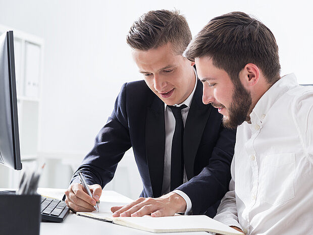 Zwei Mitarbeiter sitzen nebeneinander vor dem PC. Einer schreibt etwas in das Heft seines Kollegen und erklärt dabei.