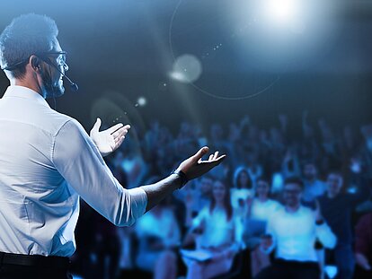 Ein Mann mit einem Mikrofon steht auf einer Bühne vor einer großen Menschenmenge