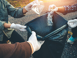 Drei Personen, jede mit einer Ecke der Mülltüte, sammeln Plastikmüll und werfen ihn in die Tüte
