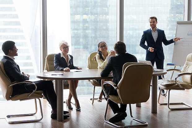 Mehrere Personen sitzen im Team-Meeting, während eine Person am Flipchart präsentiert.