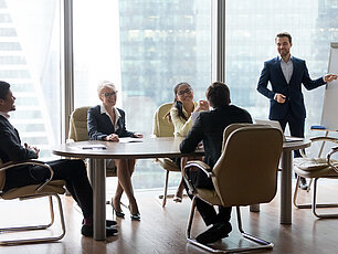 Mehrere Personen sitzen im Team-Meeting, während eine Person am Flipchart präsentiert.