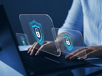 Eine Hand tippt auf einen Computer, während drei transparenten Schlössern, welche für die Cyber Security stehen, über dem Computer schweben