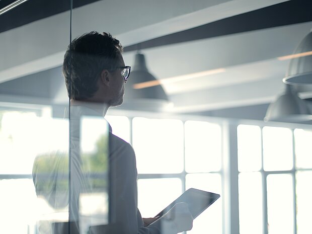 Ein Mann steht im Büro durch gläserne Türen, trägt eine Brille und hält ein iPad in der Hand, während er nach vorne schaut.