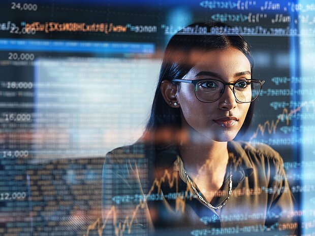Das gespiegelte Gesicht einer Frau mit Brille, auf einem Monitor der Code anzeigt.