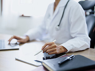 Ein Arzt sitzt in seinem Büro und arbeitet mit einem Laptop. In einer Hand ruht seine Hand auf dem Touchpad des Laptops, während die andere Hand einen Apple Pencil auf dem iPad hält, jedoch nicht schreibt.