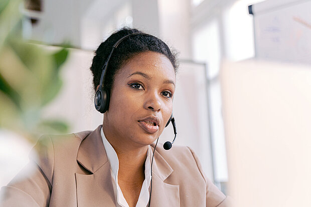 Eine Frau in braun-weißer Kleidung und mit Headset sitzt an ihrem Arbeitsplatz und führt ein Kundengespräch