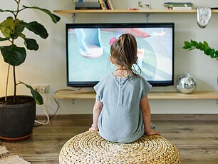 Ein Kind schaut fernsehen. Im Raum befinden sich Pflanzen und Bücher.